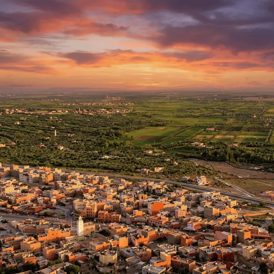 Afourar&rsquo;s dense ochre and terracotta rooftops around a white minaret at sunset, with olive groves and the flat irrigated plain stretching to distant hills